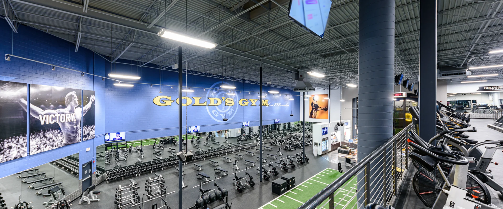 A wide-angle view of the gym layout, showcasing the free weight section, strength machines, and a functional training area. Cardio machines are positioned on an upper level, while the Gold’s Gym branding decorates the walls.