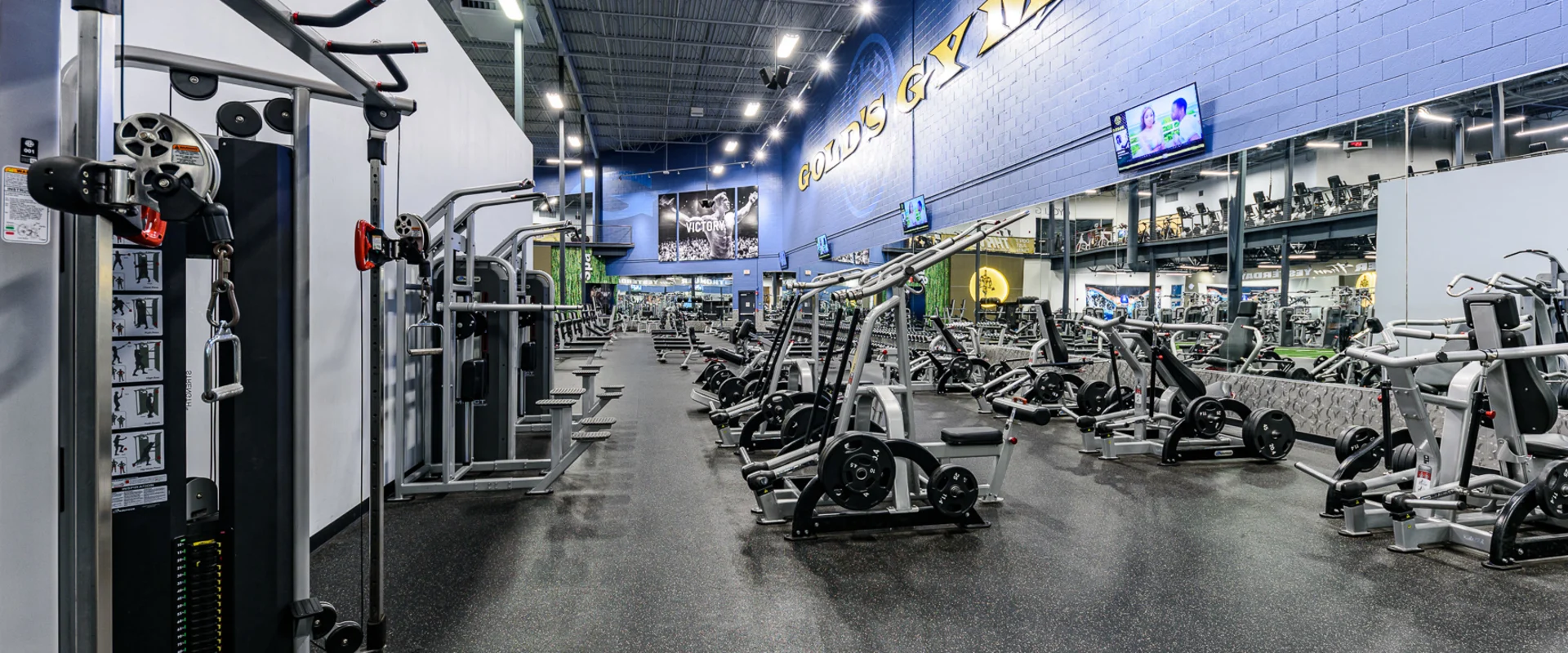 A strength training section featuring an array of plate-loaded machines, including chest press, leg press, and lat pulldown stations. The area is surrounded by gym mirrors and blue-accented walls with motivational graphics, and the machines are arranged for optimal training flow.