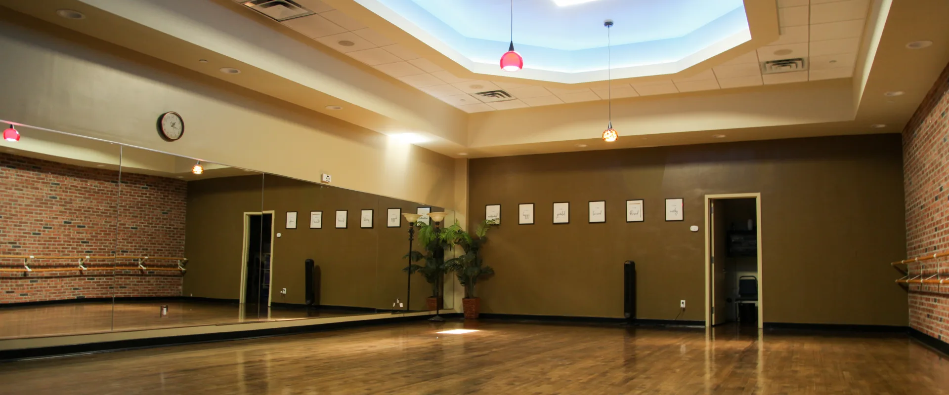 Yoga studio featuring a mirrored wall reflecting the space, an exposed brick accent wall with mounted ballet barres, polished wooden flooring, pendant lights hanging from a recessed ceiling, and framed inspirational quotes on the back wall.