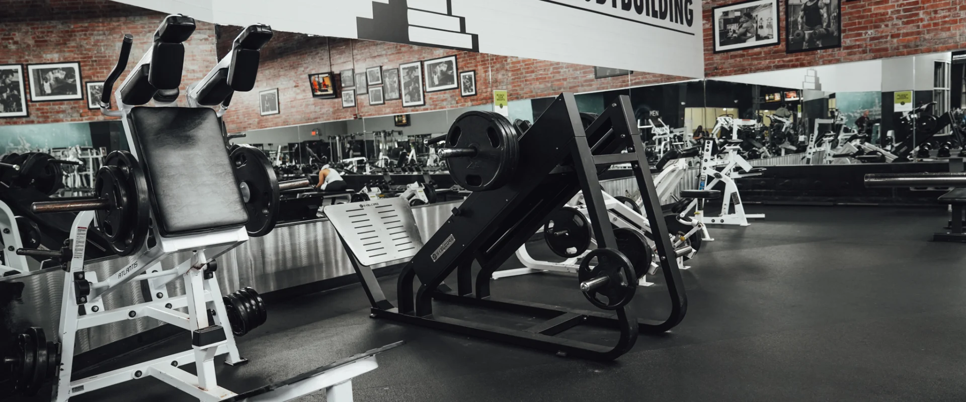Leg press and strength machines in the leg training zone at Gold’s Gym Randleman, with framed bodybuilding photographs along a brick wall and mirrored reflections of equipment.