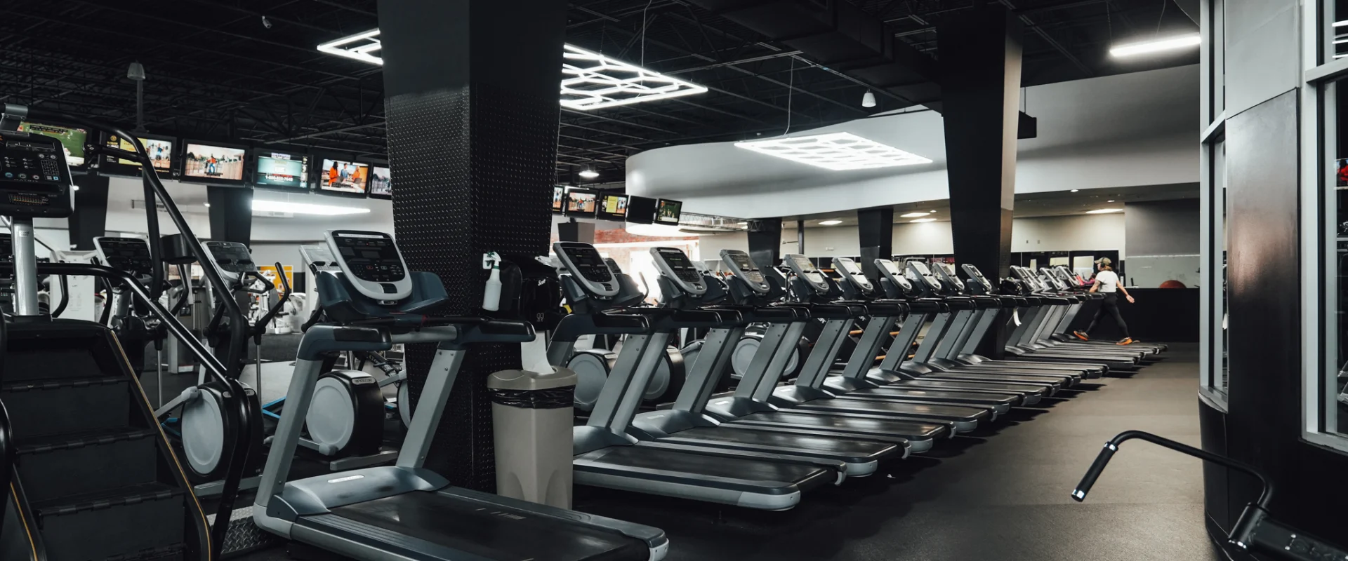 A long row of treadmills at Gold’s Gym Randleman beneath LED lighting panels, with multiple televisions mounted above.