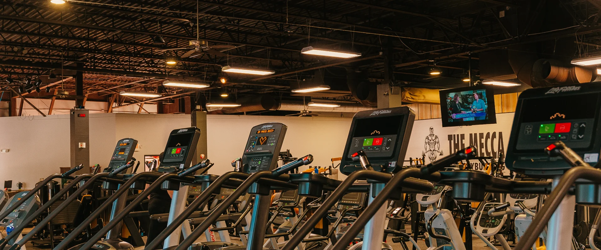 Cardio area at Gold's Gym Hendersonville, featuring a row of stair climber machines, ellipticals, and other cardio equipment. The space is illuminated by multiple lights hanging from a black ceiling, with a TV mounted on the wall for cardio-goers to watch during their workouts. The iconic Mecca logo, featuring a bodybuilder holding weights and the text "The Mecca," adds to the gym's motivating and dynamic atmosphere.