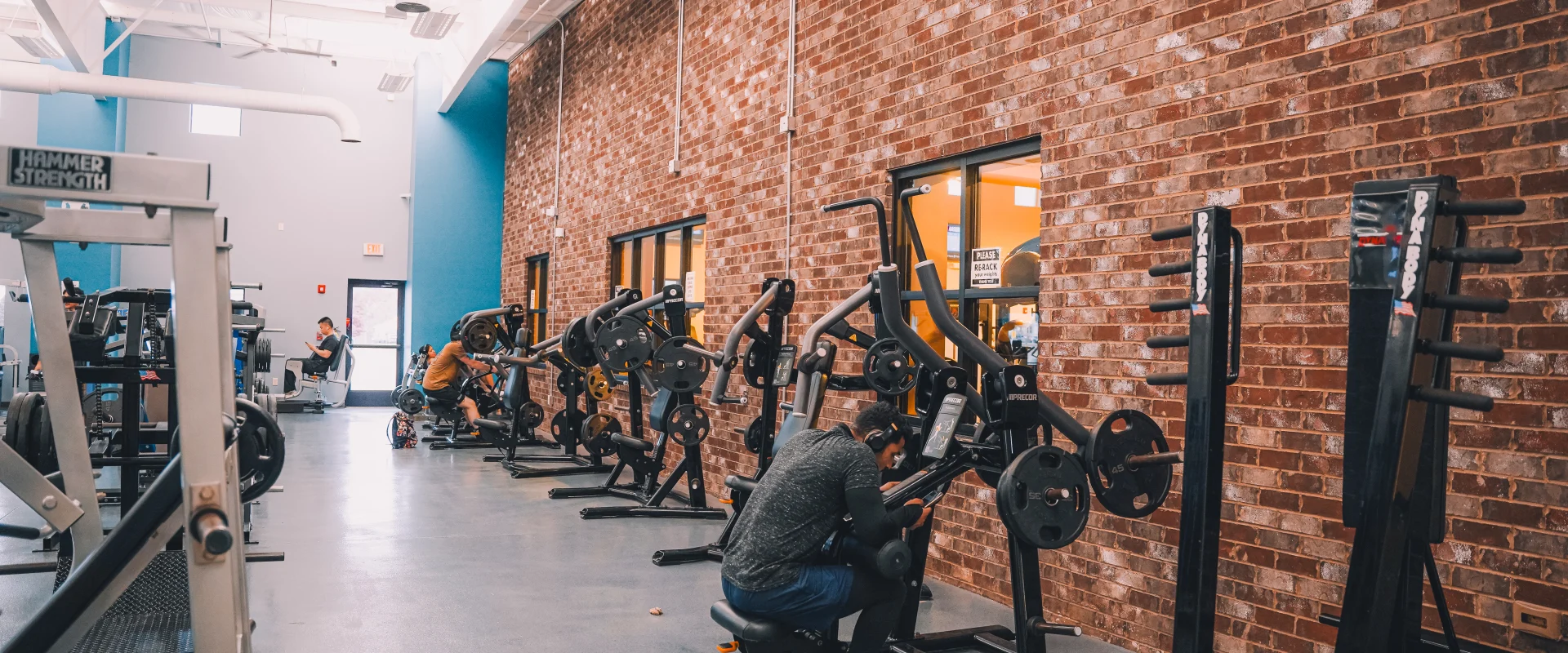 Plate-loaded machine area at Gold’s Gym Tennessee, Clarksville, featuring a row of strength training equipment set against an exposed brick wall. The space has high ceilings with exposed white ducts and hanging lights. The flooring is a polished blue-gray surface, contrasting with the black and silver machines. Windows provide a view into another area of the gym. Gym-goers are engaged in various workouts, with some using the machines and others resting nearby.