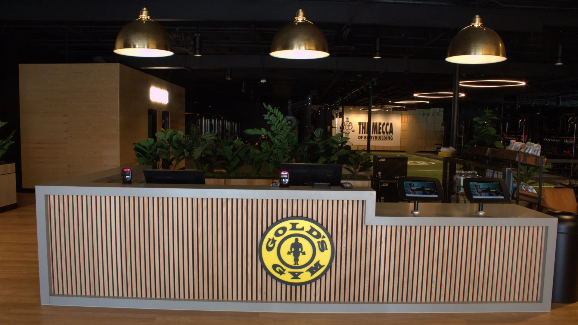 Front desk at Gold's Gym Austin, Burnet Rd featuring vertical wood slats, gold pendant lights, lush greenery, and a view of the gym floor and turf area in the background.