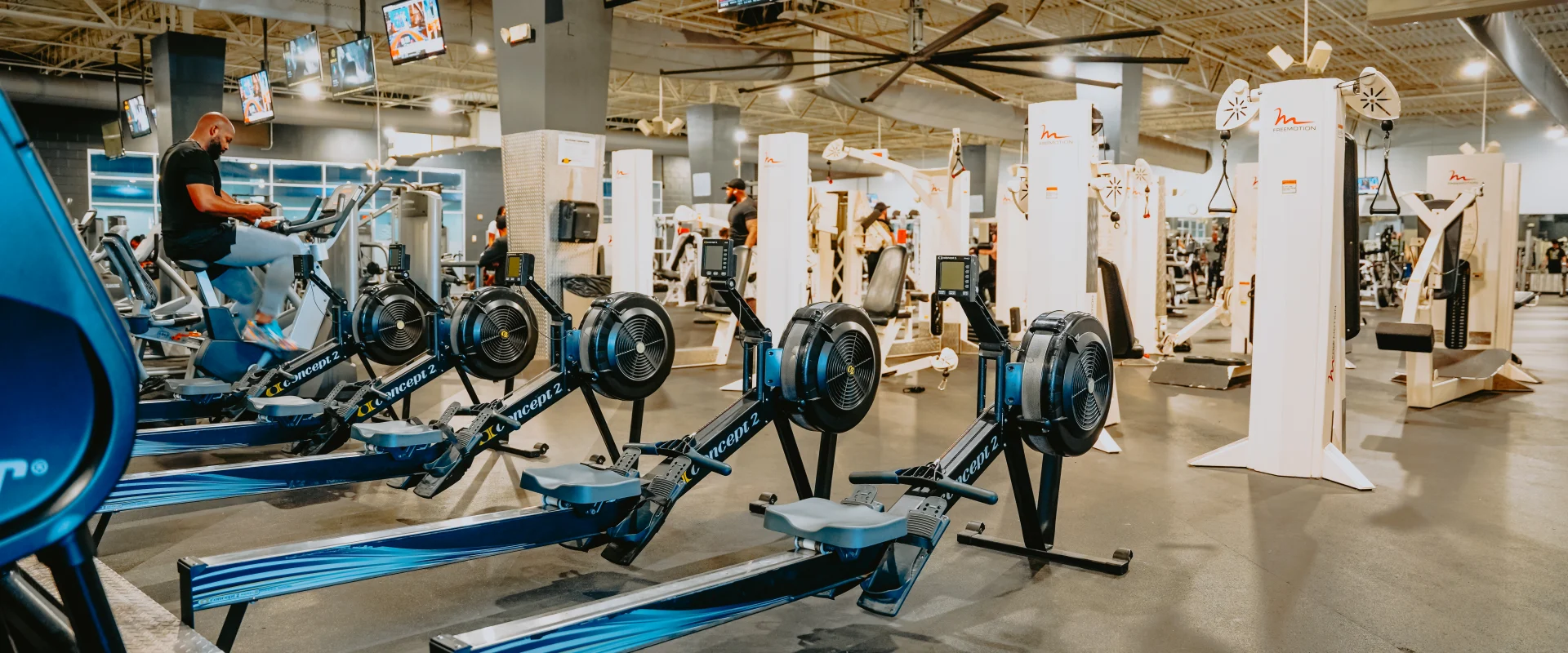 Rowing machine area at Gold’s Gym Tennessee, Lee Highway, showcasing a row of sleek rowing machines placed on a polished rubber floor. Overhead TVs display workout programs, while a high ceiling with exposed beams and ducts enhances the spacious feel. Surrounding the area are various other fitness equipment and machines, with gym members actively using the space. Bright lighting ensures the area is well-illuminated for an energetic workout environment.