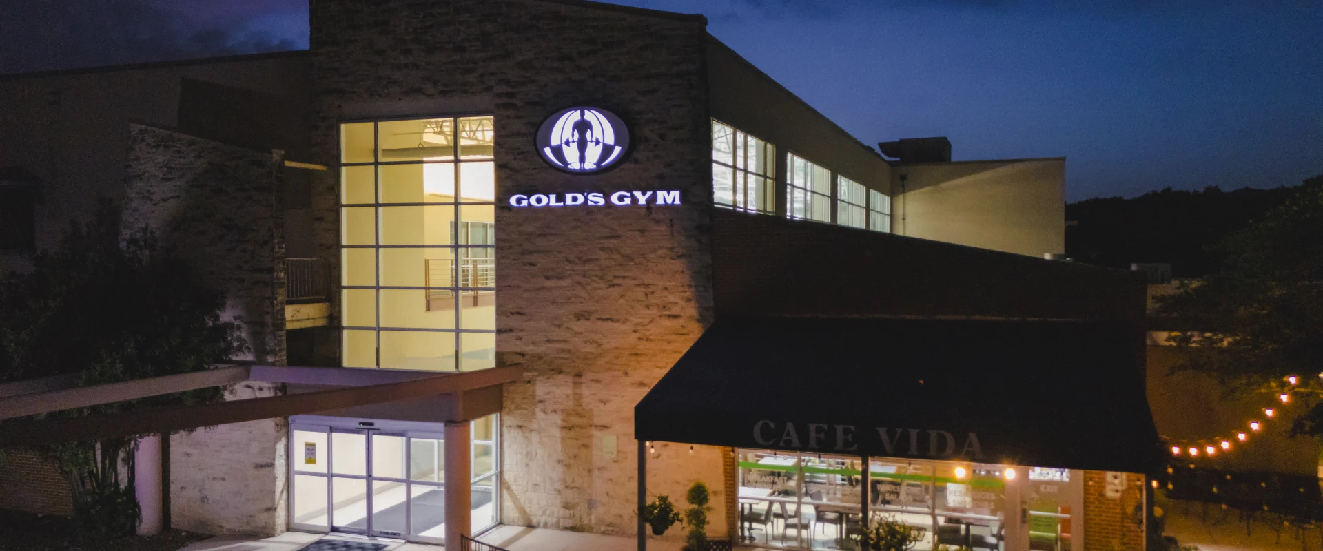 Exterior of Gold's Gym San Antonio Rogers Ranch at dusk, with the Gold's Gym logo illuminated and a neighboring café on the side of the building.