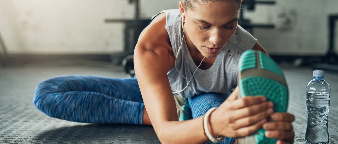 A person in a gray tank top and blue leggings is sitting on the floor, stretching their leg and using a green exercise roller.