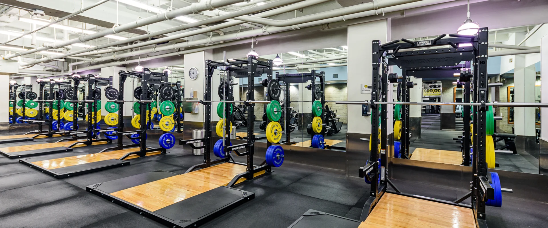 Olympic lifting platforms at Gold’s Gym Hollywood, featuring multiple black and wood-finished platforms equipped with squat racks and pull-up bars. Each station includes a barbell with color-coded bumper plates in blue, yellow, green, and black, neatly organized on storage pegs. The area is lined with floor-to-ceiling mirrors. Overhead fluorescent lights illuminate the space, with exposed pipes and ductwork visible along the ceiling. Black rubber flooring surrounds the platforms. A wall-mounted clock and instructional signage are positioned nearby.