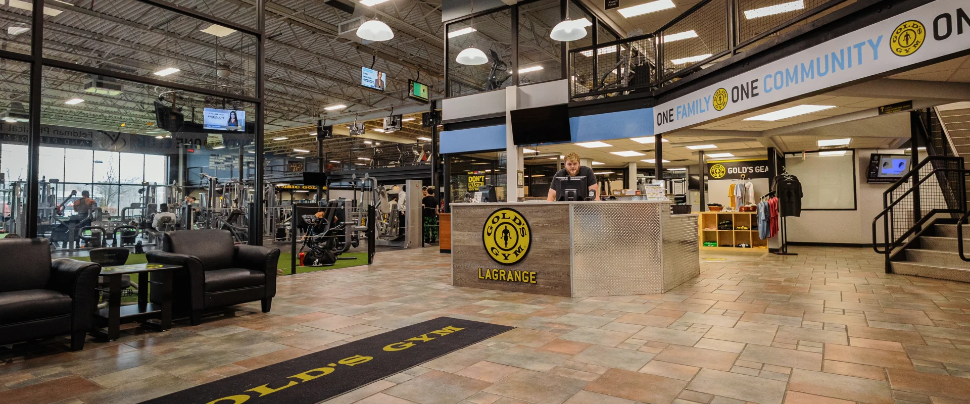 Front desk at Gold's Gym New York (Poughkeepsie), featuring a modern reception area with a wood and metal-paneled counter displaying the Gold’s Gym LaGrange logo. Pendant lights hang from the high ceiling, casting a warm glow over the tiled flooring. A lounge area with black leather chairs and a small table sits near large glass windows overlooking the gym floor, filled with strength equipment. A bold banner above the desk reads “ONE FAMILY, ONE COMMUNITY,” emphasizing the gym’s welcoming atmosphere.
