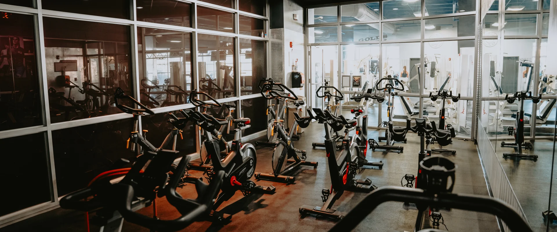 Group cycle room at Gold’s Gym Tennessee, Downtown Chattanooga, featuring rows of stationary bikes on a dark rubber floor. The space includes a mirrored wall, glass partitions, and overhead lighting. A hallway and adjacent workout area with weight equipment are visible through the glass, creating a bright and open atmosphere.