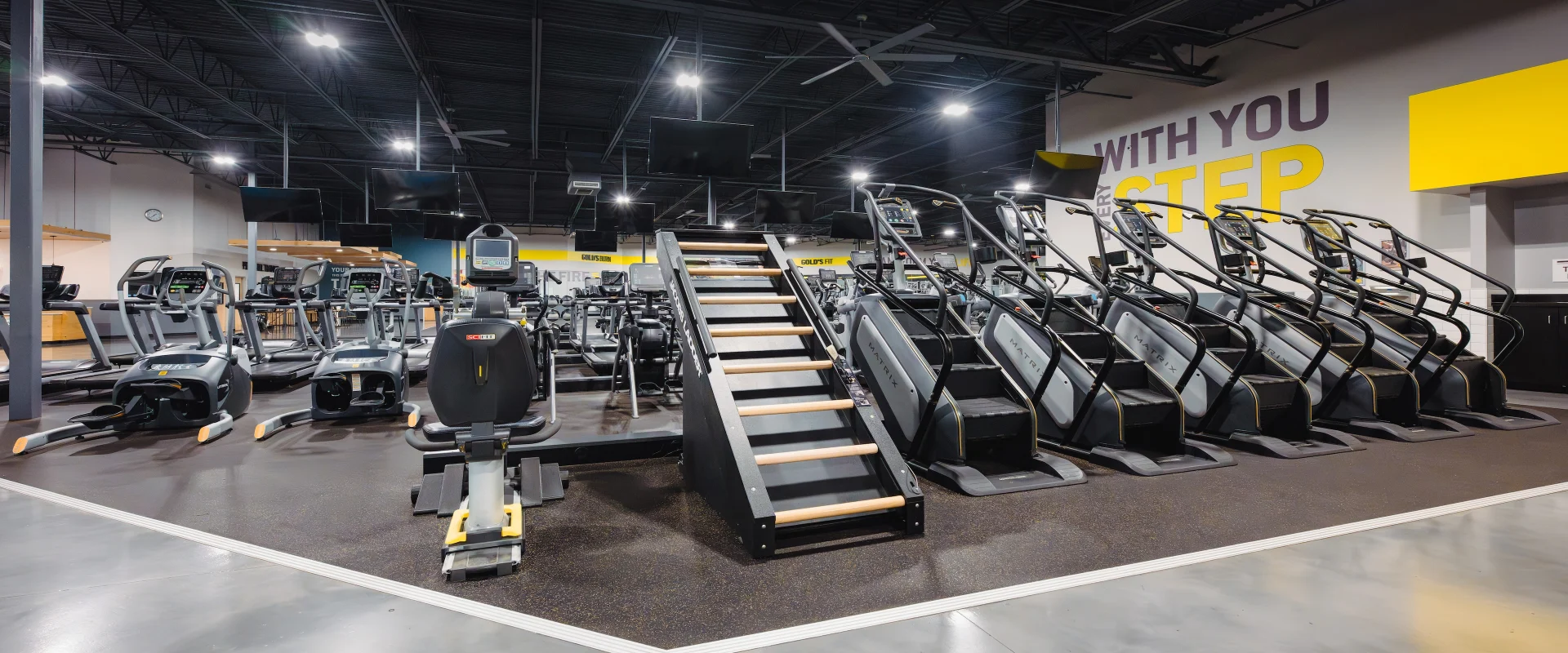 The cardio area at Gold's Gym in Texas (Slaughter & South Congress) features a variety of equipment, including treadmills, stair climbers, ellipticals, and rowers. The space is well-lit with overhead lighting, and flat-screen TVs are mounted for entertainment. The black ceiling with exposed ducts contrasts with the bright workout area below.