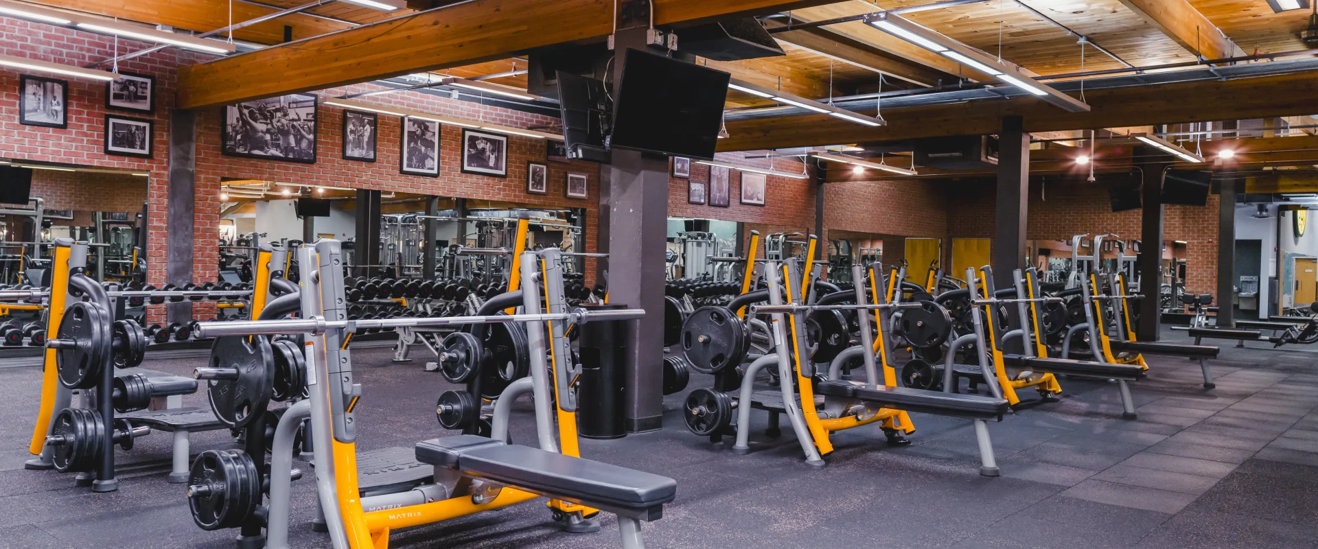 A gym section with multiple weightlifting benches and racks holding barbell plates, set against a brick wall with framed photos and a wooden ceiling.