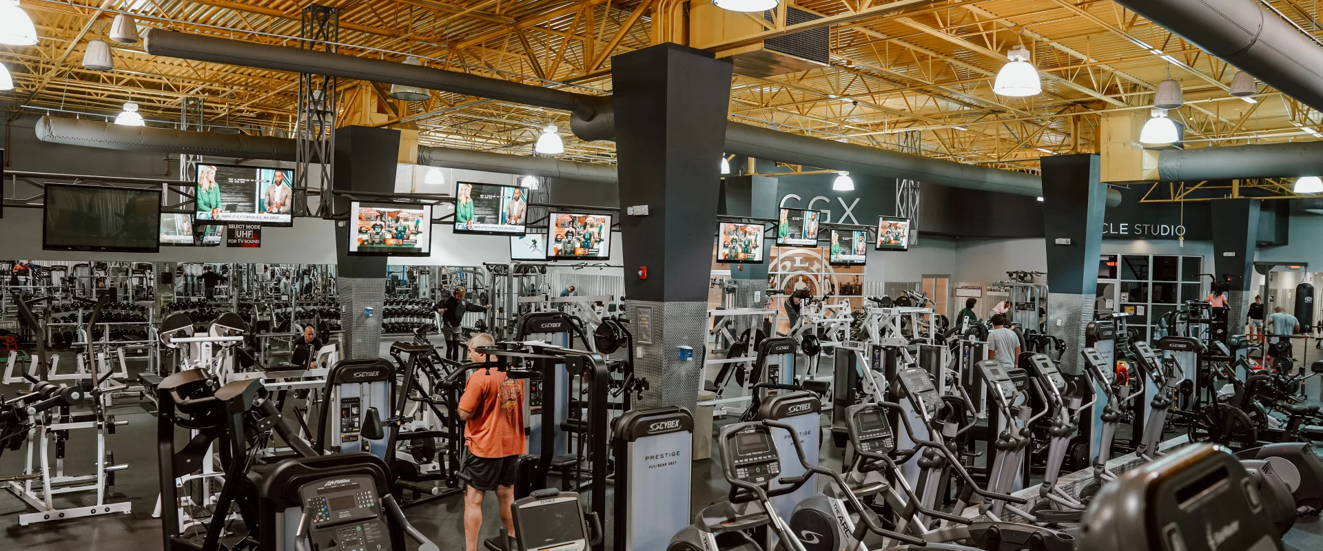 Members working out in the cardio and machine weights area at Gold’s Gym Lenoir City, Tennessee. The gym features an array of treadmills, elliptical machines, and weight-training equipment arranged in rows. Overhead, yellow industrial-style ceiling beams and hanging lights illuminate the space, while multiple mounted TV screens display entertainment for gym-goers. The open layout and mirrored walls create a spacious and energetic workout environment.