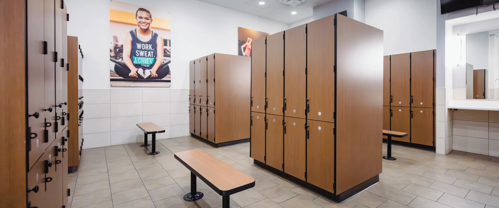 Locker Rooms at Gold’s Gym in Texas (N. Round Rock), featuring rows of wooden lockers with black metal hinges and built-in locks. The space includes tiled flooring and benches with wooden tops and black metal legs placed between the locker rows for seating. Bright overhead lighting illuminates the area, complemented by wall-mounted mirrors near the sink area. Motivational fitness posters adorn the walls, adding a vibrant and energetic atmosphere to the clean and organized layout.