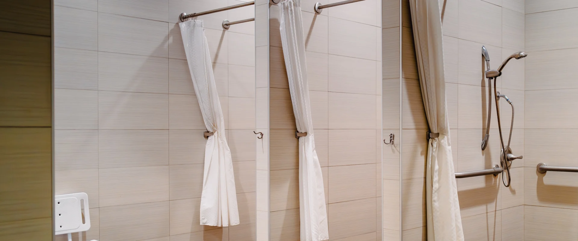 Accessible shower stalls in a clean facility, featuring light beige tiles, white curtains, and grab bars. A white shower chair is visible on the left.