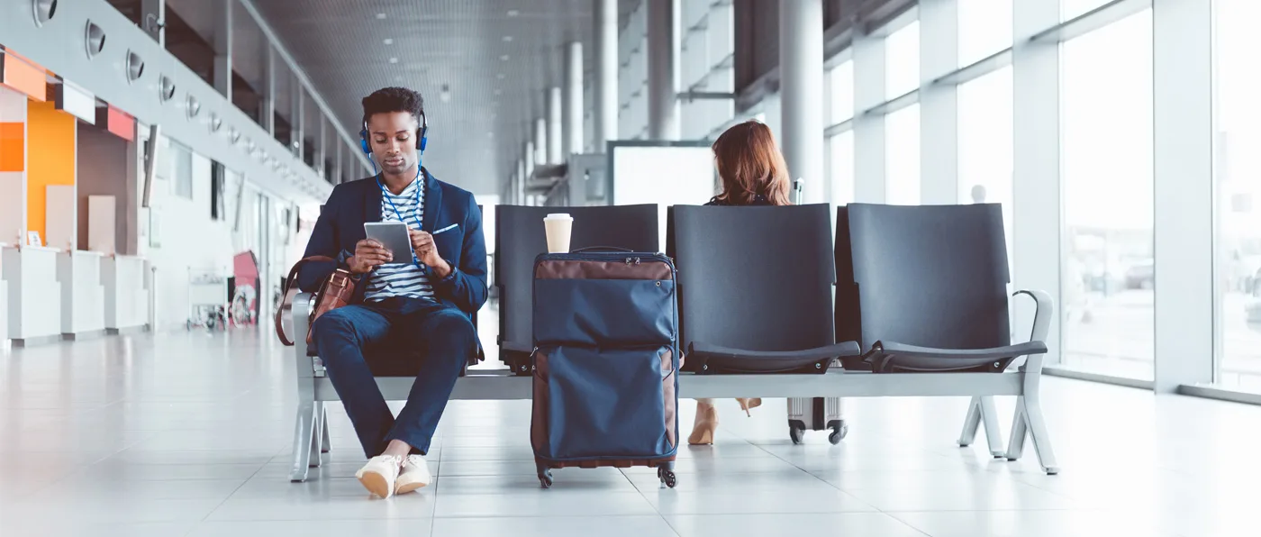 The image shows a person sitting on a chair in an airport terminal, with another person standing nearby and luggage nearby.