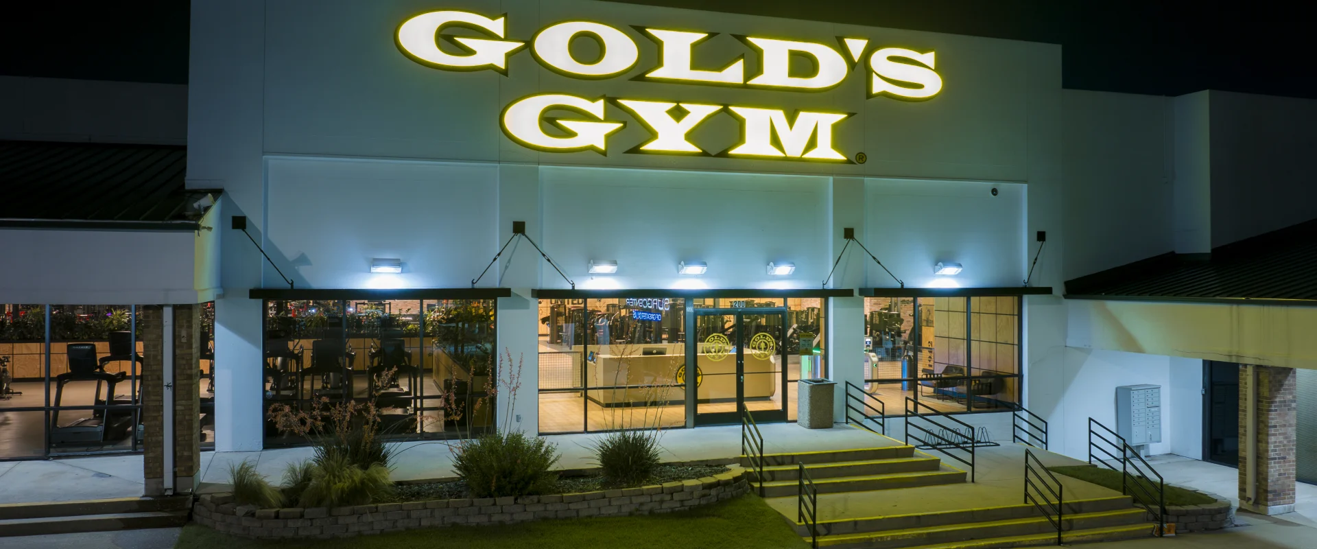 Nighttime exterior of Gold’s Gym Highland Meadows with a brightly illuminated gold sign above the entrance. The modern facade features large glass doors and windows showcasing the interior. The surrounding area includes landscaped greenery, accessible ramps, and a parking lot in the foreground, all softly lit by overhead lights.