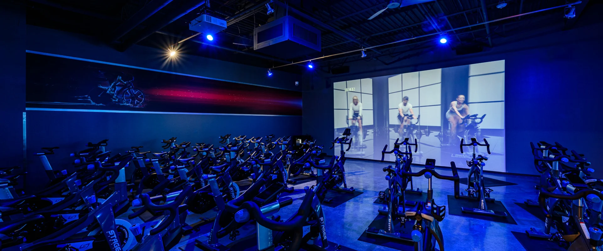 Gold’s Gym Thousand Oaks immersive group cycle studio with rows of stationary bikes facing a large projection screen displaying a virtual cycling class. Blue LED lighting highlights the dark industrial-style ceiling, which features exposed ductwork and ceiling fans. A futuristic cycling-themed mural runs along the side wall. The polished concrete floor reflects the lighting, enhancing the high-energy atmosphere of the studio.