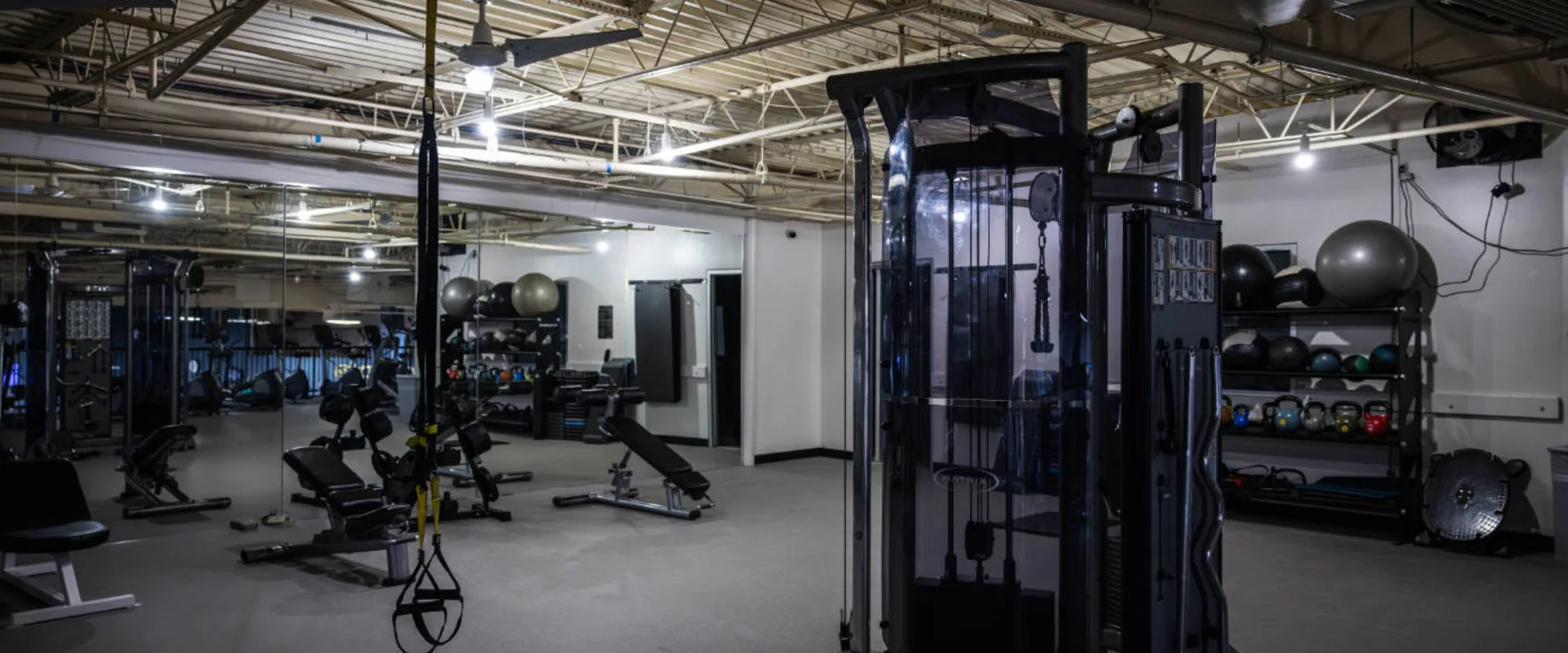 A functional training area at Gold’s Gym in Woodbridge (Lake Ridge) features a cable machine with multiple attachments, a TRX suspension trainer hanging from the ceiling, and adjustable benches positioned near a mirrored wall. A rack of kettlebells, stability balls, and medicine balls lines the back wall, offering a variety of training options. Overhead industrial-style lighting illuminates the space, while the open layout provides ample room for dynamic workouts.