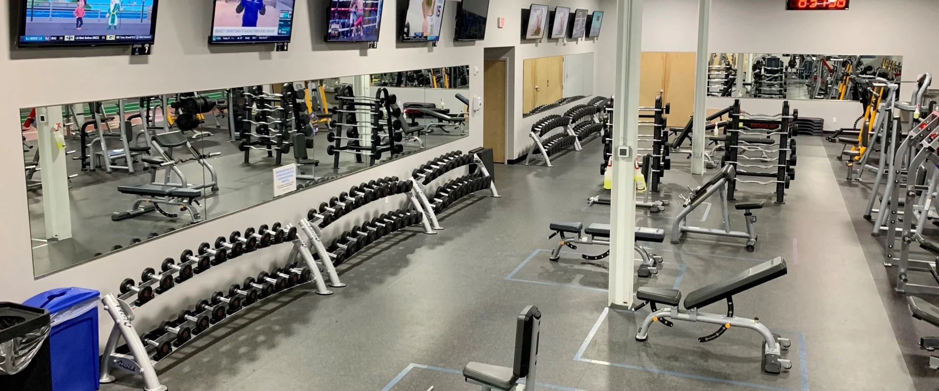 A spacious and well-equipped gym with various exercise equipment, including weight racks, benches, and cardio machines, displayed on the walls and throughout the room.