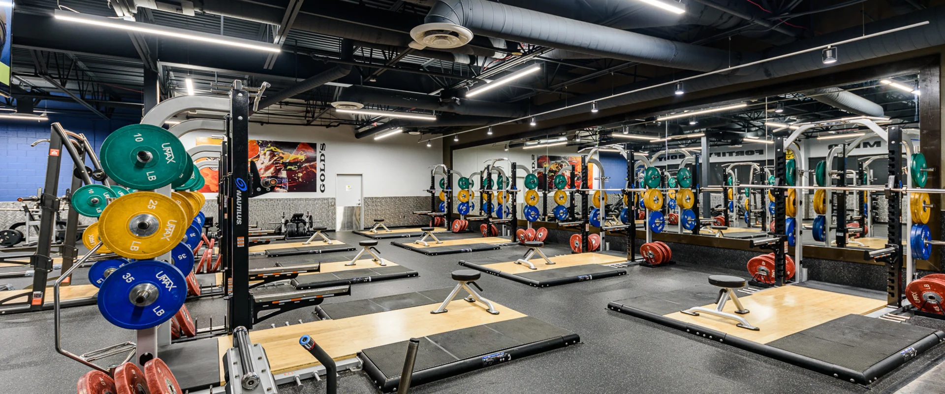 A spacious Olympic lifting area equipped with multiple wooden lifting platforms, heavy-duty squat racks, and barbells with color-coded weight plates. Mirrors line the back wall, reflecting additional weightlifting equipment and workout areas. Overhead industrial lighting provides bright illumination.