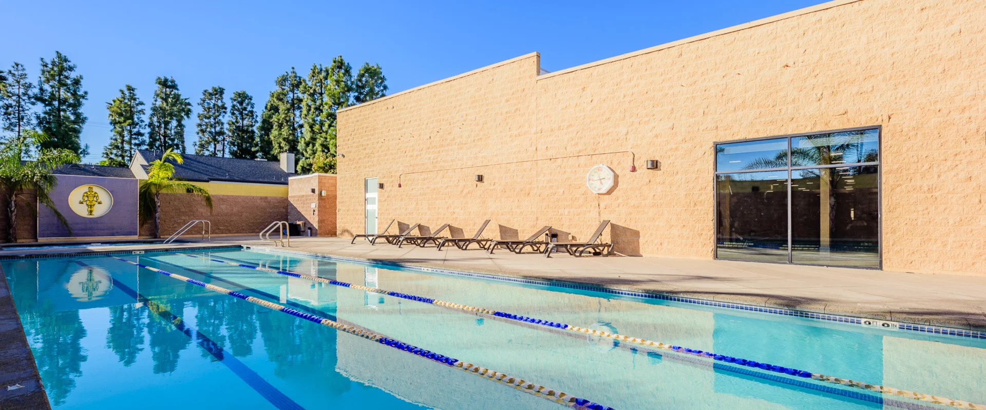 Outdoor swimming pool at Gold's Gym Garden Grove, featuring multiple lanes marked by blue and yellow floating dividers. The pool deck is lined with lounge chairs, and a privacy wall with a large Gold’s Gym logo adds branding. Tall trees and palm trees surround the area, with clear blue skies reflecting in the water.