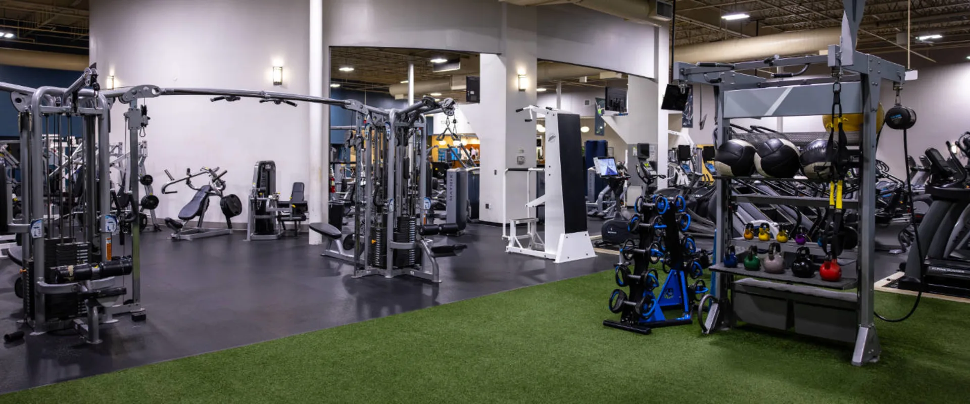 Turf training area at Gold’s Gym in Woodbridge (Lake Ridge), featuring a dedicated functional fitness space with kettlebells, medicine balls, TRX suspension trainers, and dumbbells. Adjacent weight machines and cable stations provide additional strength training options. The gym's sleek, modern design includes bright lighting and open workout spaces.