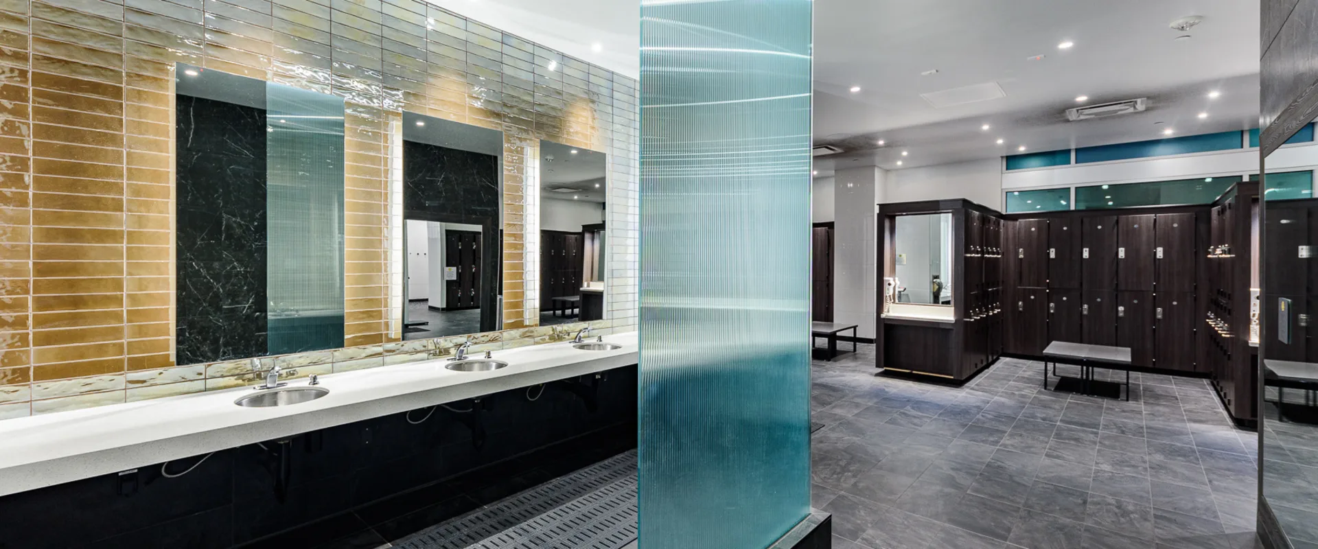 Locker room at Gold’s Gym Long Beach, featuring dark wood lockers, sleek tiled floors, and well-lit vanity stations. A modern glass partition separates the sink area, which includes a long countertop with multiple stainless steel basins and large mirrors. Benches provide seating, creating a spacious and functional environment for members to freshen up before and after workouts.