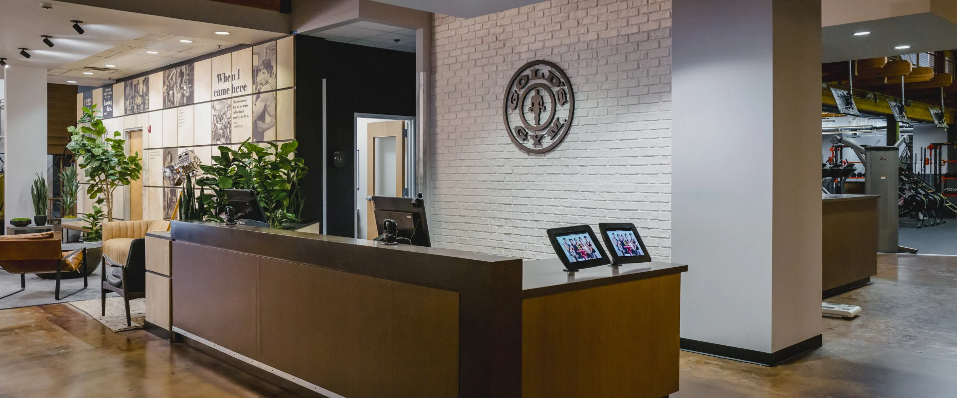 A modern front desk at Gold's Gym with a sleek wooden counter, white brick wall, and greenery, creating a welcoming and professional atmosphere.