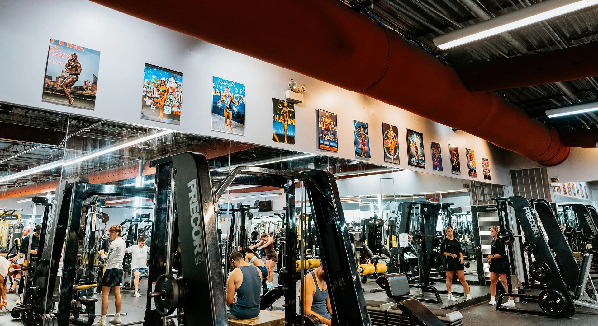 Members exercising in the plate-loaded equipment area at Gold’s Gym Bellevue, Nashville, Tennessee. The gym features machines and members engaged in various exercises. The mirrored wall reflects the equipment and gym-goers, while a series of bodybuilding competition posters and awards line the upper wall. Overhead, industrial-style lighting and a large red ceiling duct add to the gym's dynamic ambiance.