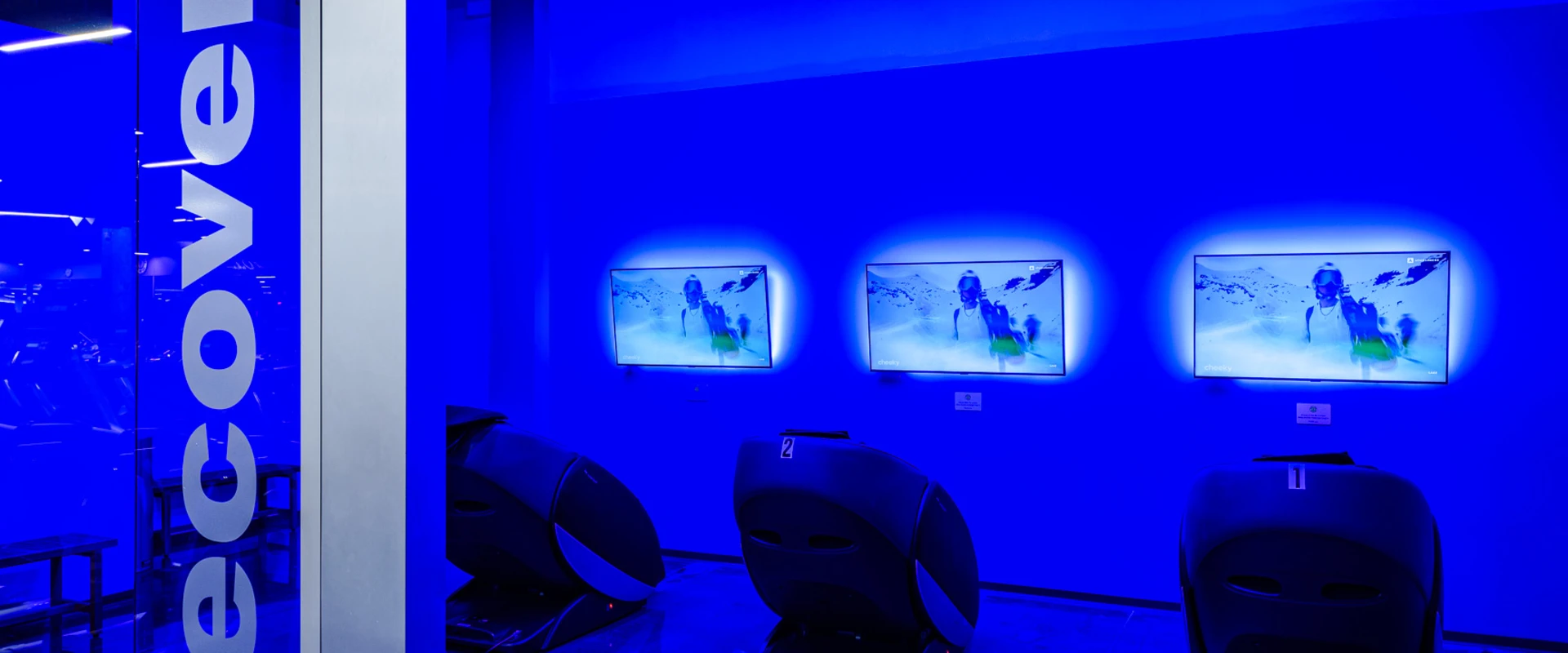 Recovery area at Gold’s Gym Thousand Oaks, designed for post-workout relaxation and muscle recovery. The space features multiple hydro massage chairs, each facing a mounted screen displaying calming visuals. Blue ambient lighting creates a tranquil atmosphere. The glass entrance is labeled "Recovery," emphasizing the area’s focus on rest and rejuvenation.