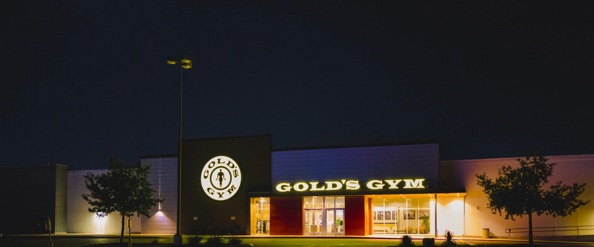Exterior of Gold's Gym San Marcos at night, illuminated by yellow signage and lighting. The building features a large Gold's Gym logo on the facade and trees framing the entrance, with a bright moon shining in the night sky.