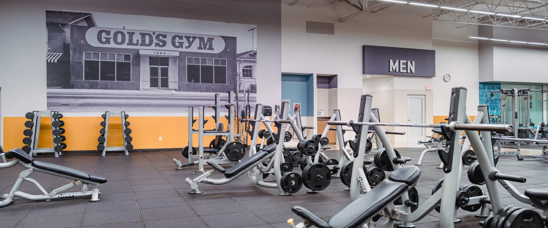 Free weights area at Gold's Gym in Texas (Pflugerville), showcasing multiple benches, weight racks, and barbells neatly arranged on rubber flooring. A large black-and-white mural of the original Gold's Gym adds a historic touch, with the men's locker room entrance visible in the background under bright, open lighting.