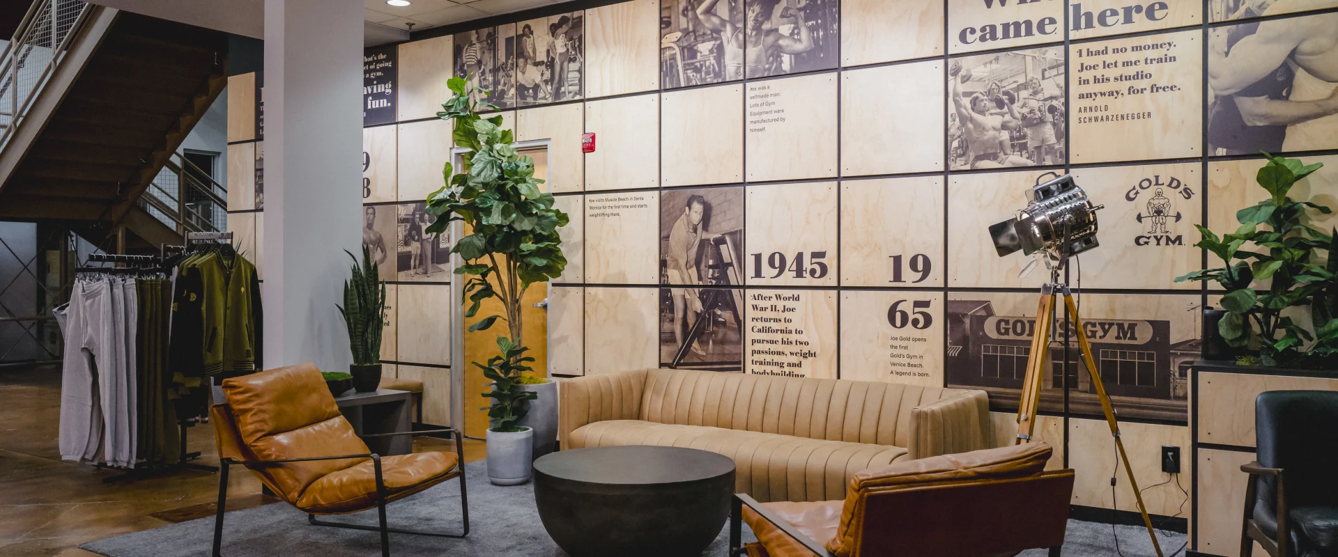 A stylish lounge area at Gold's Gym with a tan couch, leather chairs, a rug, greenery, and a wall featuring historical Gold's Gym photos and milestones.