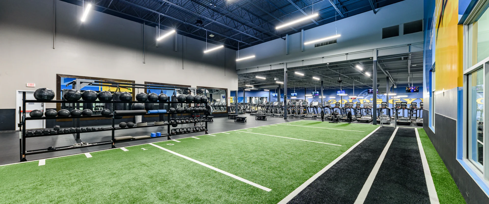 A large indoor turf training zone with sprint lanes, agility drill sections, and resistance sled tracks. The open-concept space includes a black rubber track running parallel to the turf, along with additional functional training tools like kettlebells and battle ropes.