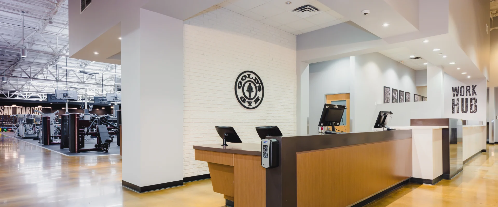 Front desk area at Gold's Gym San Marcos with a white brick wall, Gold's Gym logo, polished floors, and modern workstations creating a welcoming environment.