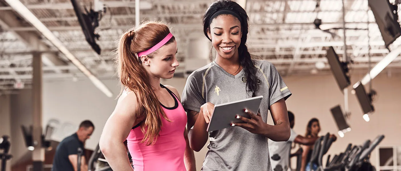 Two young women, one with long red hair and the other with dark hair, are standing together and smiling as they look at a tablet device in the woman with dark hair's hands. The background appears to be a gym or fitness center, with exercise equipment and other people visible in the background.