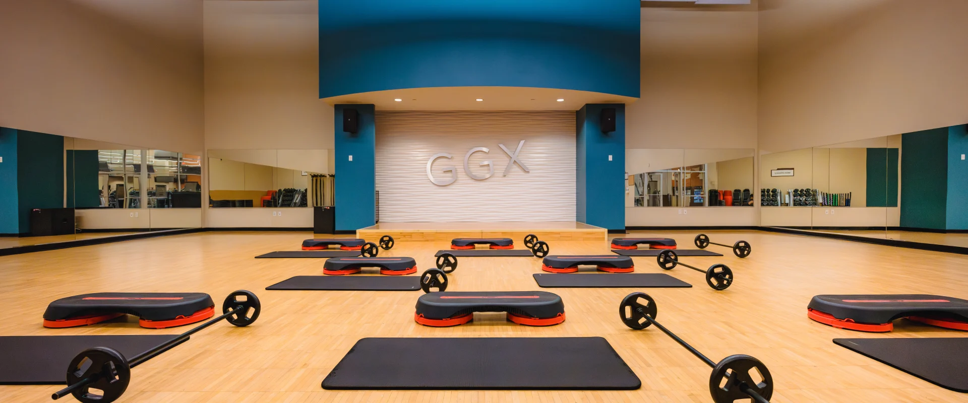 Group exercise studio at Gold’s Gym Austin (Anderson Arbor) featuring a wooden floor, step platforms, mats, and barbells arranged in rows. A stage with the “GGX” logo is centered on the teal and white accent wall, with large mirrors lining the sides of the room.
