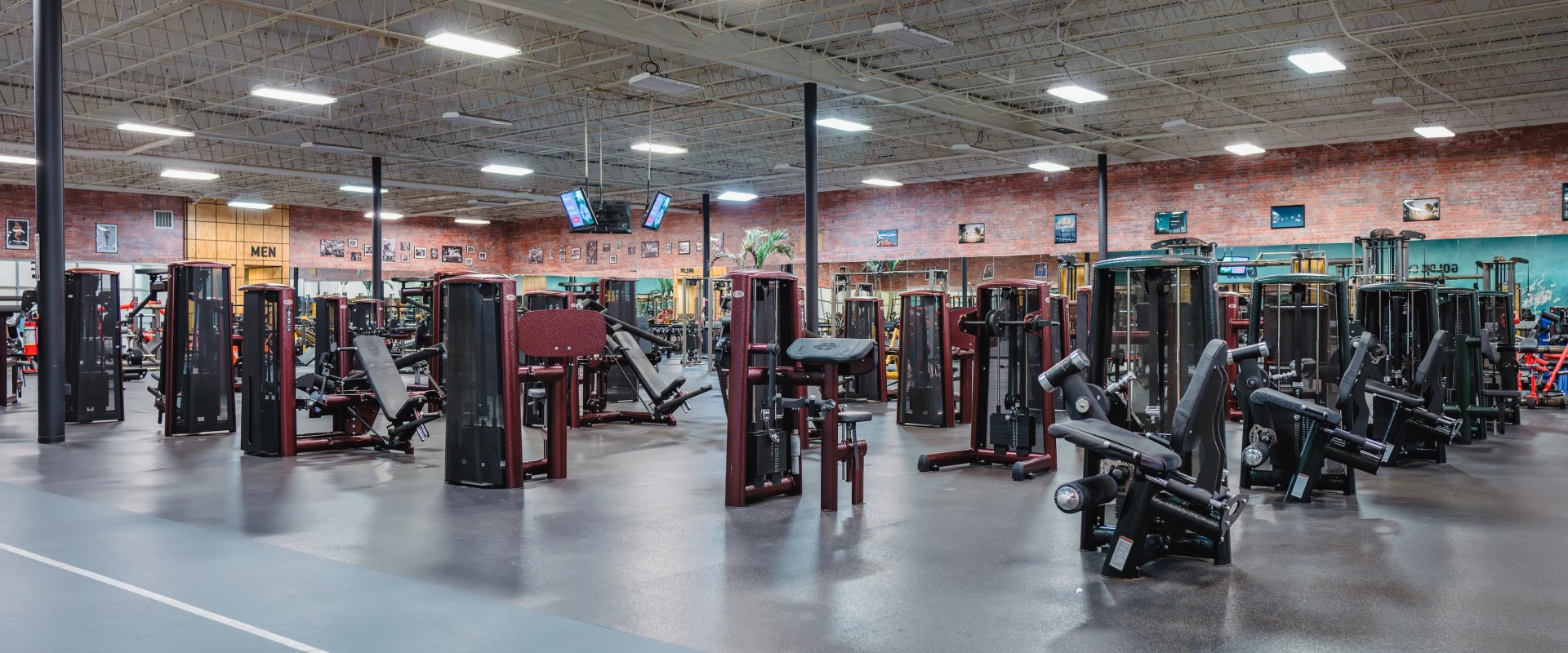 Spacious gym interior with rows of modern exercise machines on a polished floor. High ceiling with bright lights creates an open, inviting atmosphere.