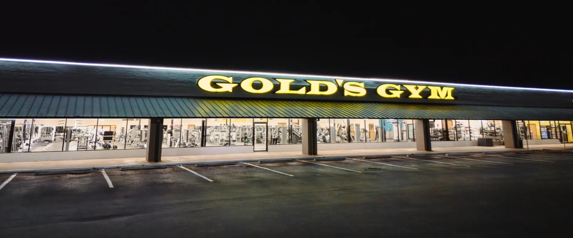Exterior view of Gold’s Gym in Austin (Southeast) at night, with the gym’s bright yellow signage illuminated against the dark sky. Large front windows reveal a glimpse of the gym’s interior, while an empty parking lot stretches across the foreground.