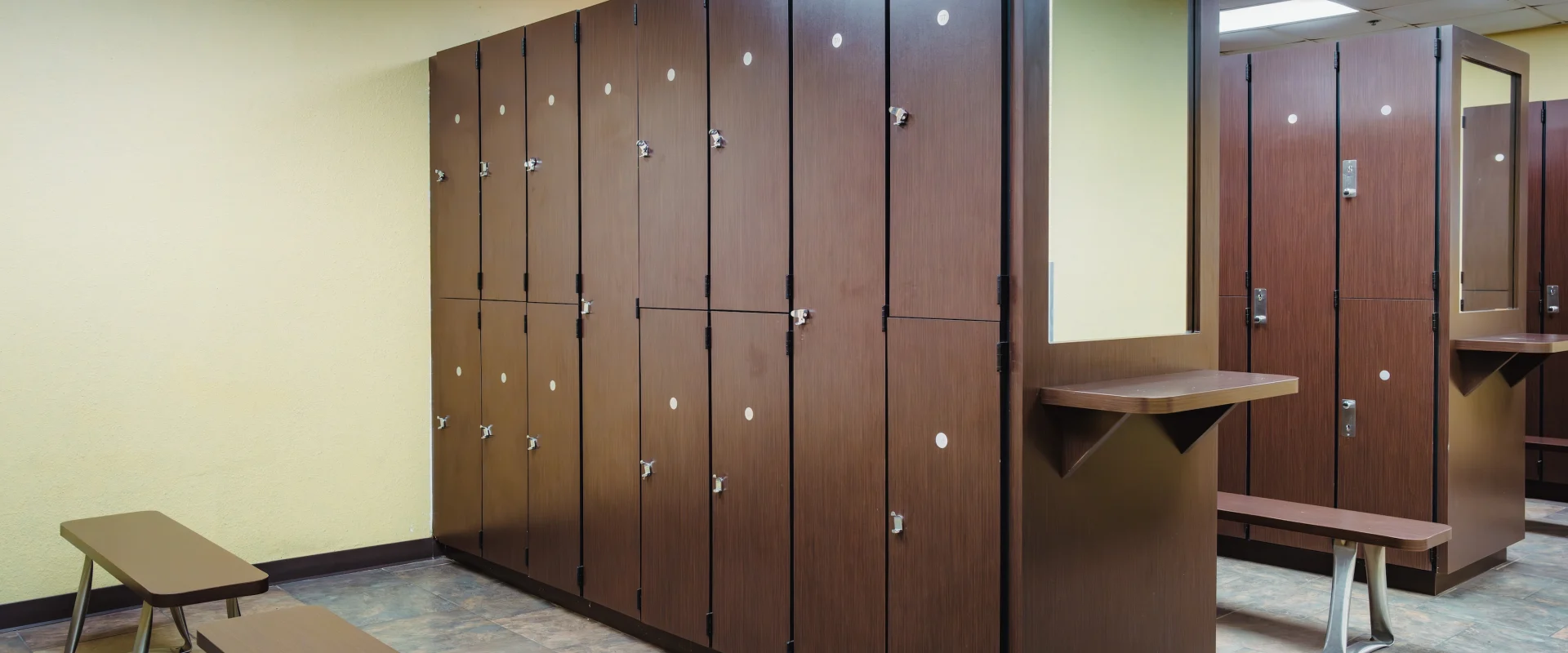 Spacious locker room with brown lockers, wooden benches, and a beige tiled floor. The setting is clean and organized, creating a calm and inviting atmosphere.