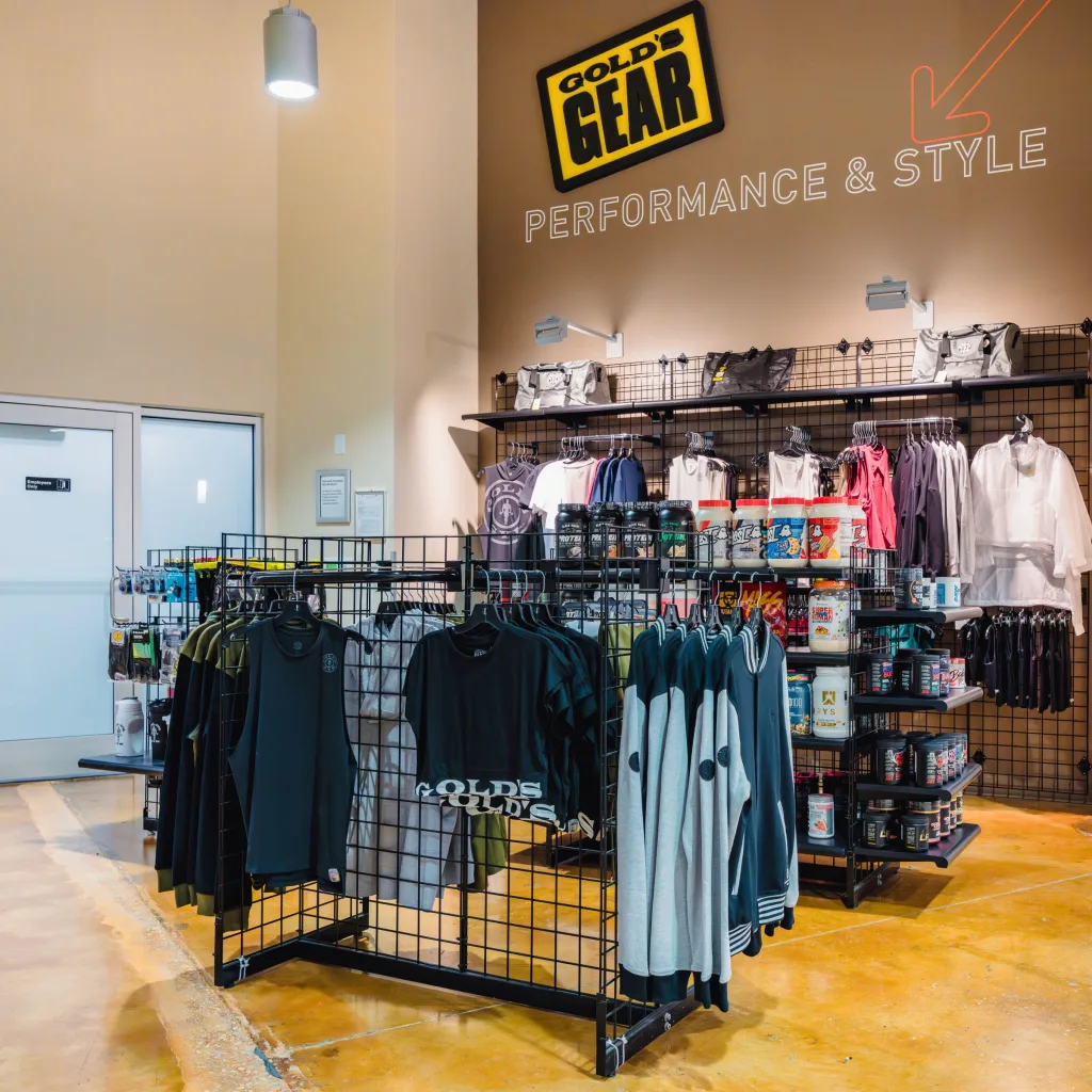 Gym retail display with racks of Gold’s Gym clothing, shelves of supplements, and a wall sign reading “Gold’s Gear Performance & Style,” set in a clean, open lobby area.
