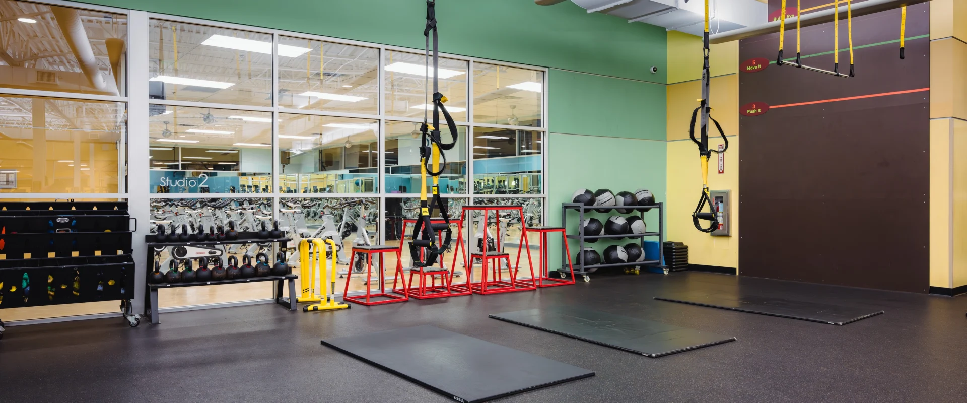 Spacious gym area with black mats on the floor, TRX straps hanging from the ceiling, kettlebells, red plyometric boxes, and large windows, creating an energetic atmosphere.