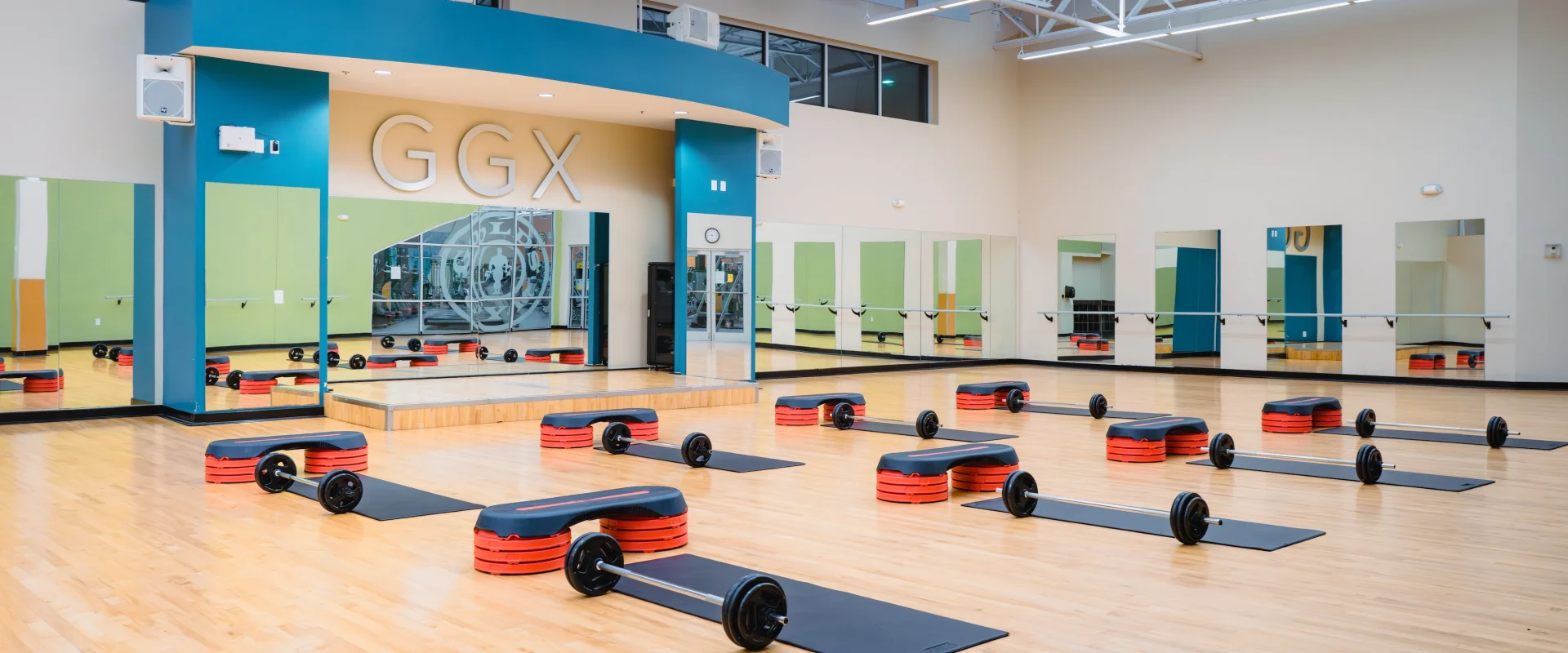 Spacious gym studio with wood floors, mirrored walls, and "GGX" sign. Weight bars and step platforms arranged neatly, ready for a class.