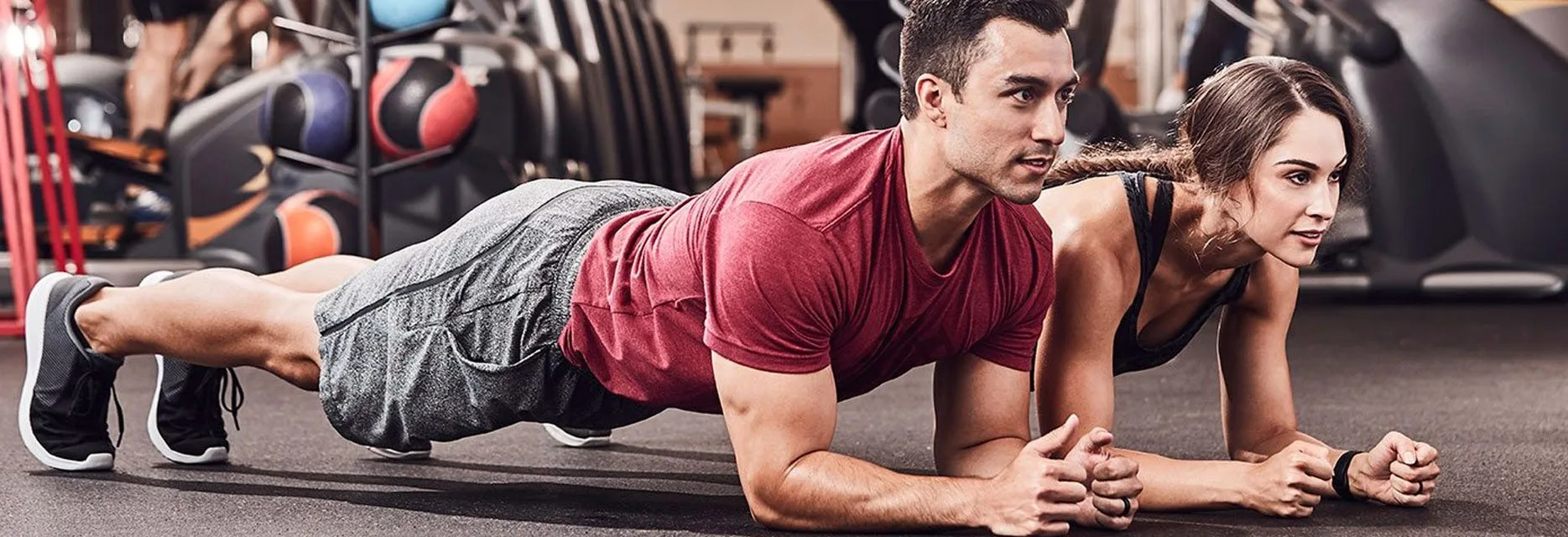 Two people, a man and a woman, are engaged in a plank exercise on a gym floor surrounded by exercise equipment.