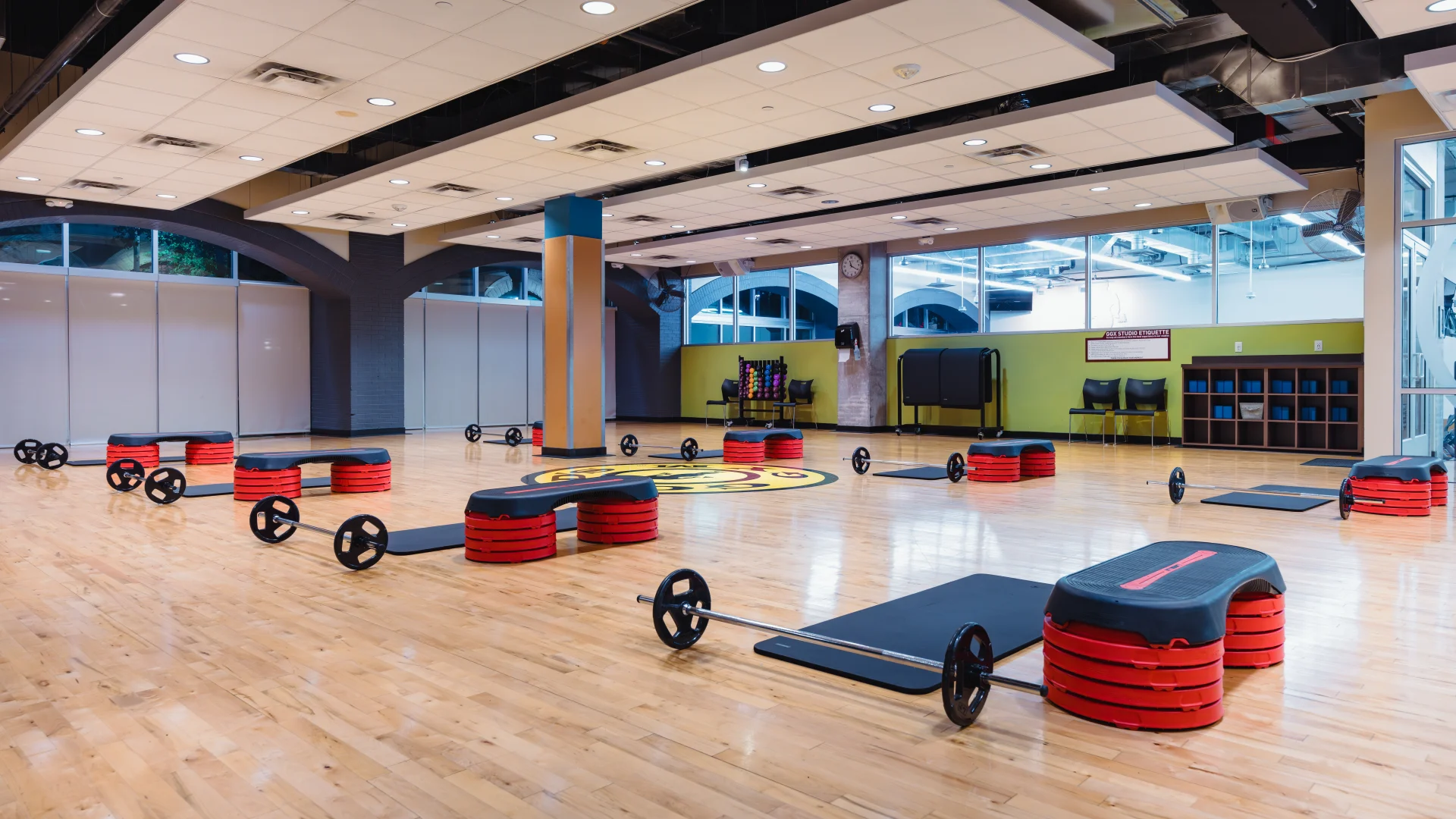 Group exercise studio at Gold's Gym Austin (Downtown) with polished wooden floors, floor-to-ceiling windows, and mirrored walls. The room is equipped with barbell sets, step platforms, and yoga mats arranged for a class.