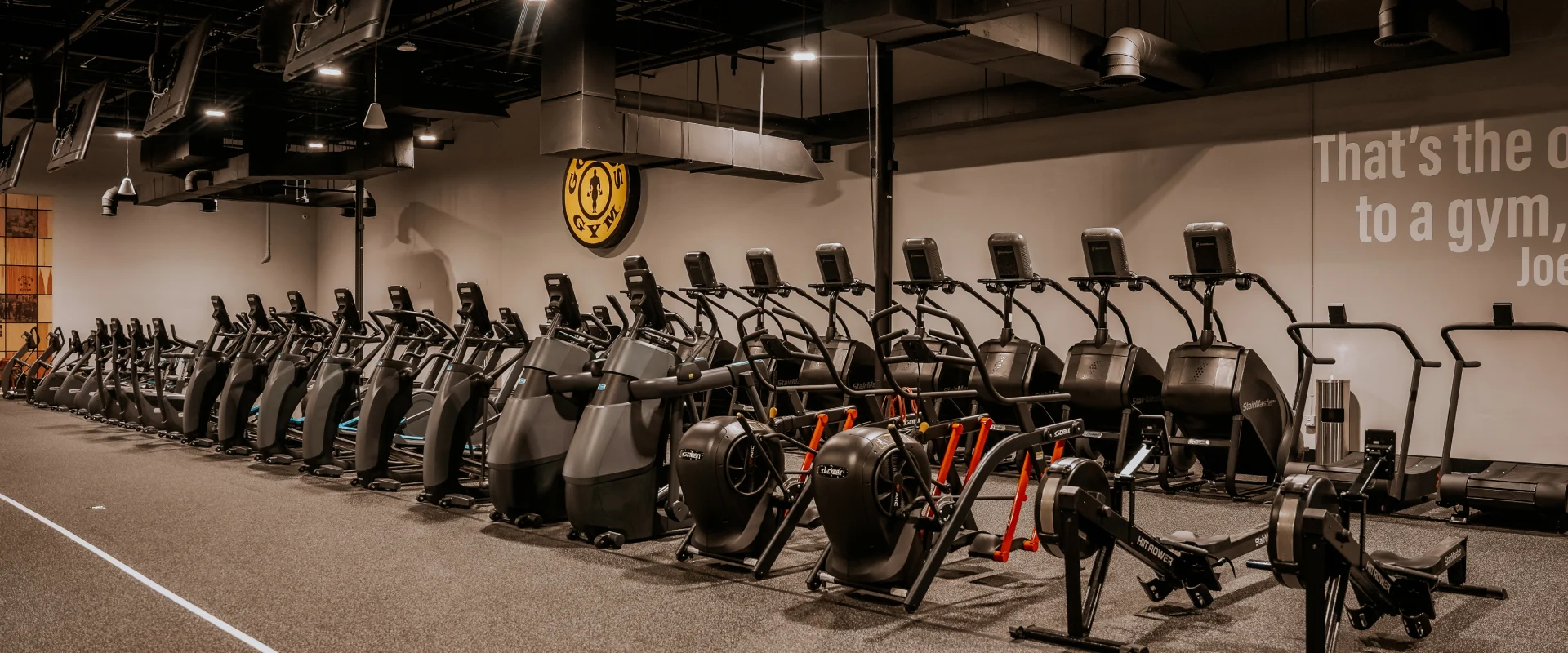 Gold's Gym Memphis, Tennessee (White Station) cardio area featuring a row of treadmills, ellipticals, and stair climbers positioned against a wall with motivational text. Several air bikes and rowing machines are arranged in the foreground. Overhead TVs line the ceiling, providing entertainment for gym-goers. The space has a dark industrial-style ceiling with exposed ductwork, and a Gold's Gym logo is mounted on the wall. The flooring includes a designated track area for functional cardio workouts.