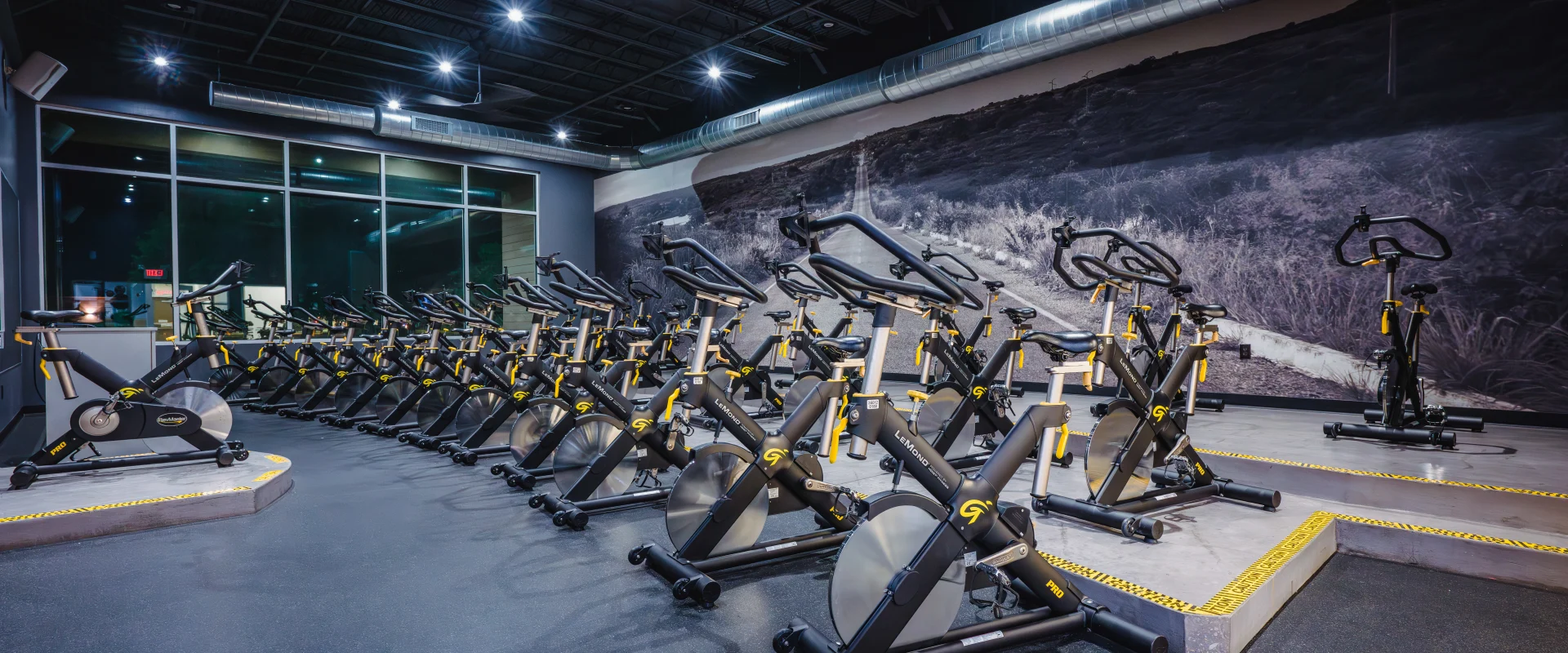 Group Cycle studio at Gold's Gym Oklahoma City (Moore) featuring tiered rows of black stationary bikes with yellow accents, a scenic wall mural, and a windowed exterior wall illuminated by overhead lighting.