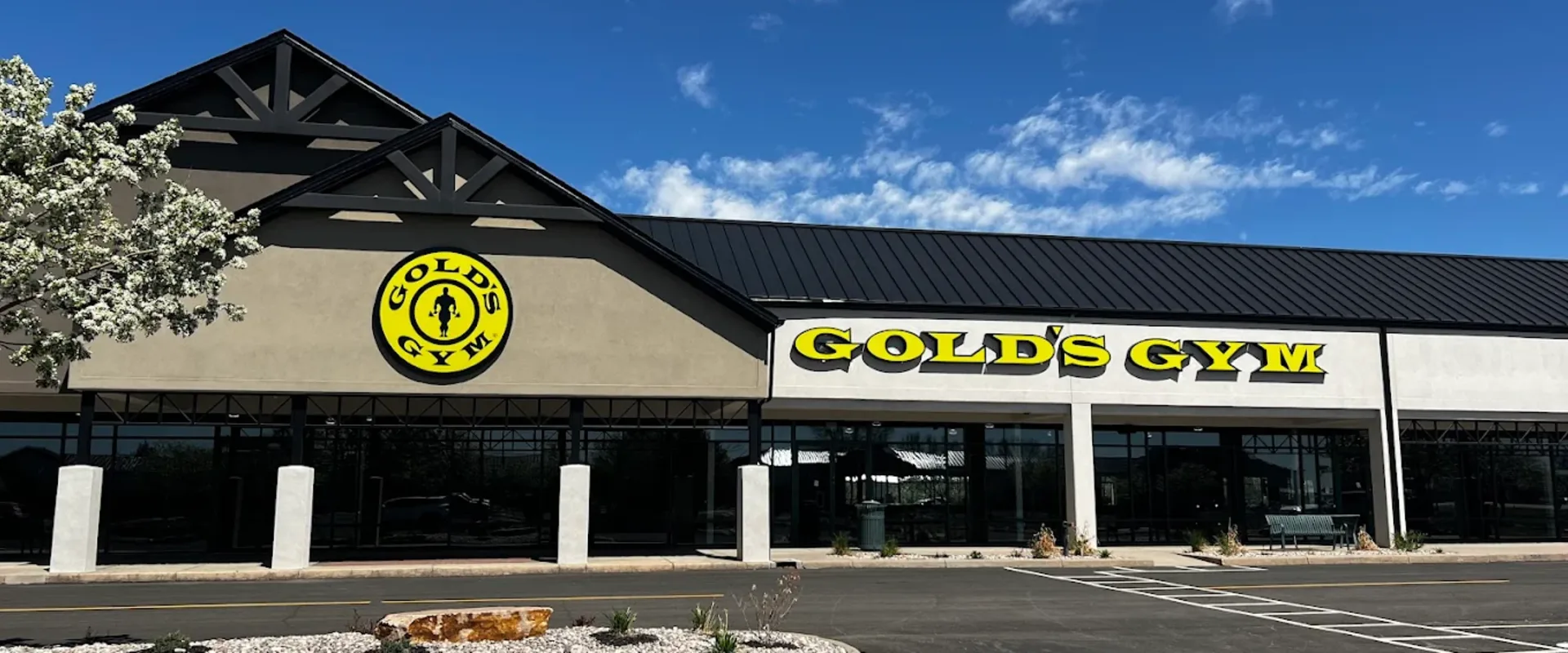 Exterior of Gold’s Gym Loveland with beige facade, black roof, and bright yellow logo on a sunny day.