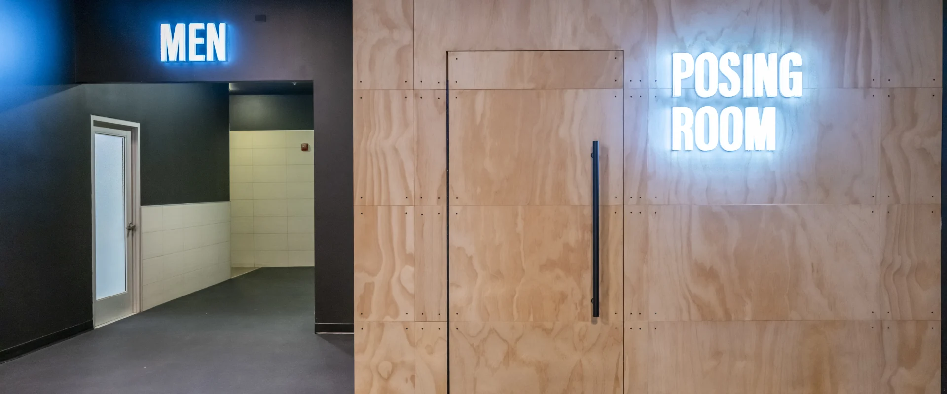 A hallway at Gold's Gym with a sign for the men's locker room and a wooden wall featuring a glowing 'Posing Room' sign, creating a modern gym vibe.
